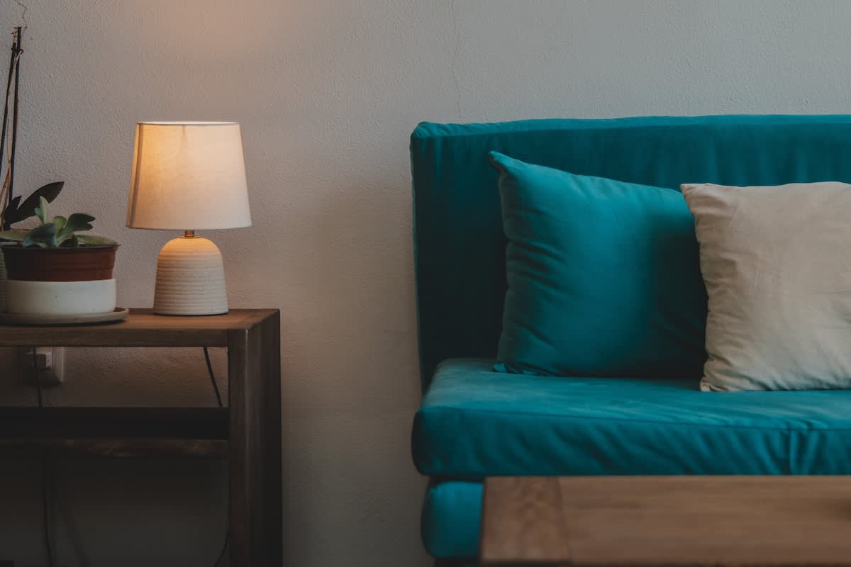 A cozy living room corner features a vibrant teal sofa with a matching pillow, a wooden side table holding a lit ceramic lamp, and a small potted plant, set against a light-colored wall.
