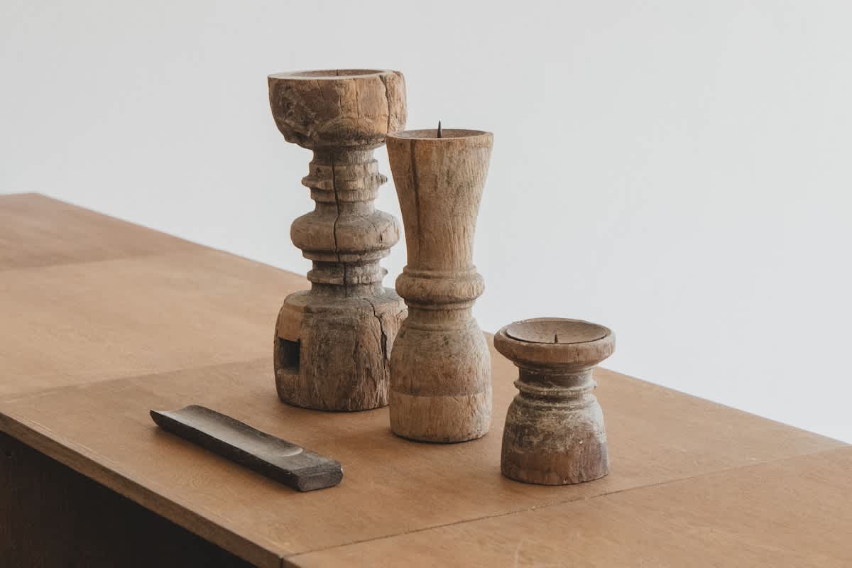 Three rustic wooden candle holders of varying heights and a small wooden tray are artfully displayed on a light-colored wooden surface against a plain white wall. The weathered, natural wood finish of the decor pieces adds a touch of organic charm.