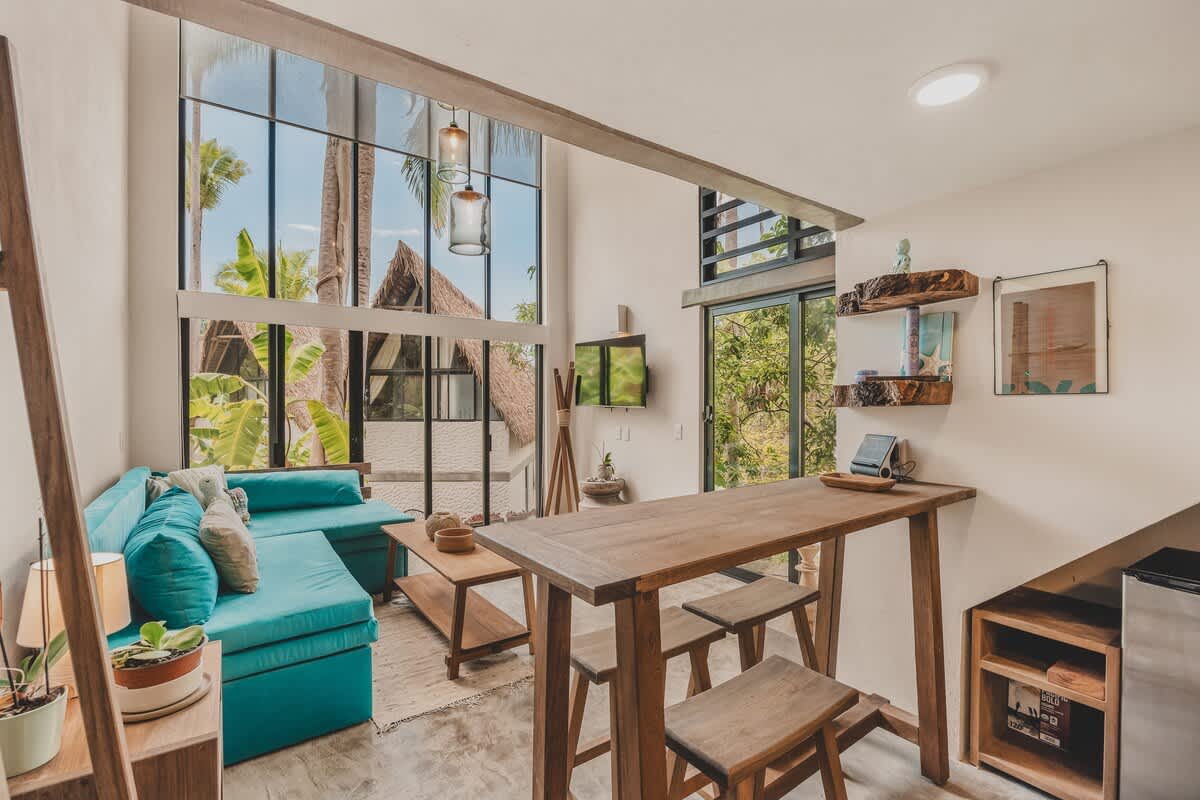 Living area featuring a teal sectional sofa, a wooden coffee table, and a high wooden dining table with stools. Large windows offer views of the tropical surroundings, and a wall-mounted TV is visible.
