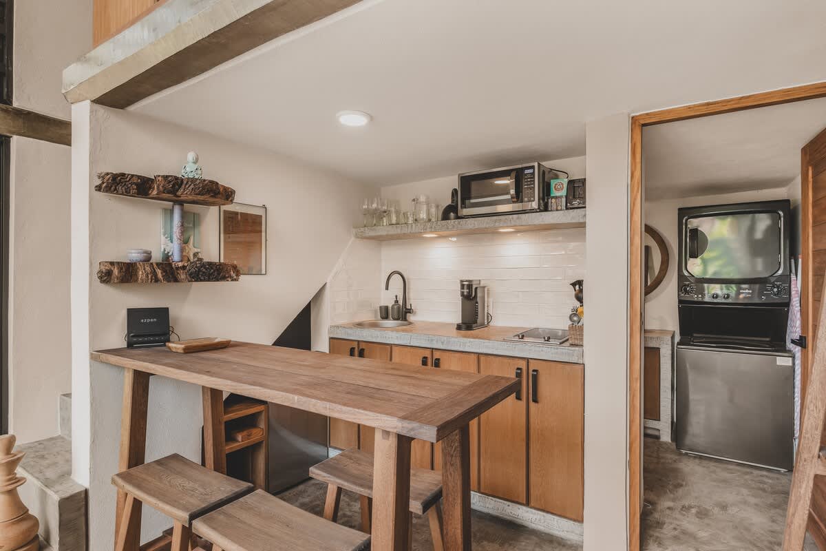Modern kitchen area with a wooden breakfast bar and stools, concrete countertops, and wooden cabinets. It features a microwave, coffee maker, and a small refrigerator, with a laundry area visible through an open doorway.