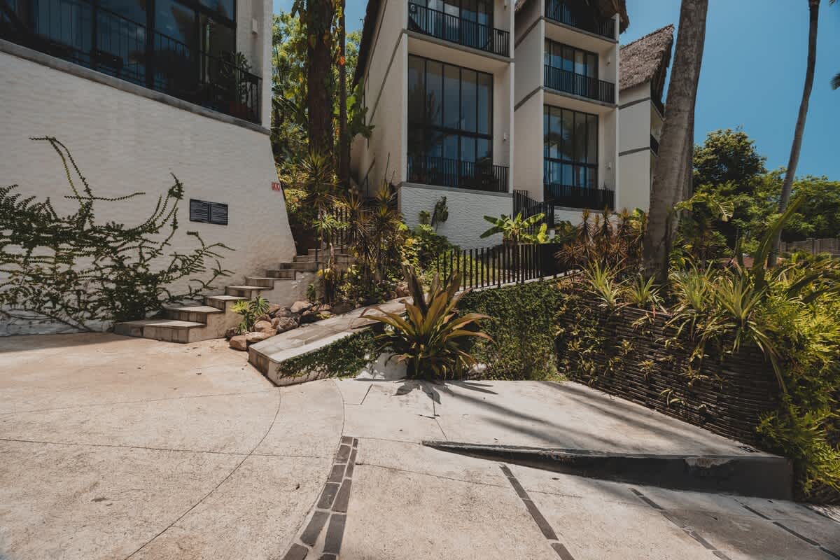 A concrete parking area with a drain leads to a set of steps ascending towards modern buildings, surrounded by lush green foliage and a retaining wall.