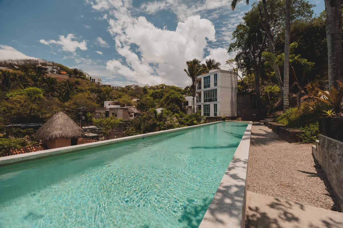 A long, narrow swimming pool with light blue water, bordered by a concrete edge and gravel, set against a backdrop of lush green hills and distant buildings under a partly cloudy sky.