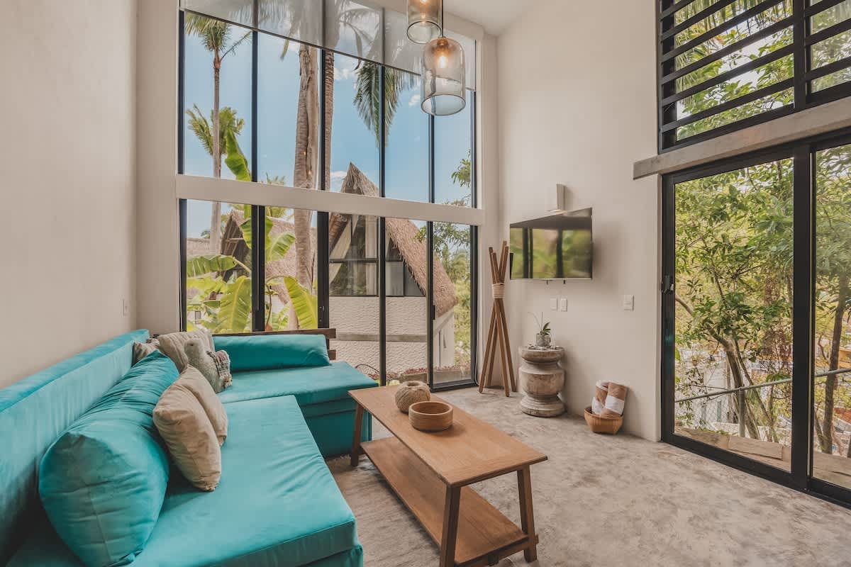 Spacious living room with a teal sectional sofa, a wooden coffee table, and a wall-mounted TV. Large windows and a sliding glass door provide abundant natural light and views of the surrounding jungle and a thatched-roof structure.