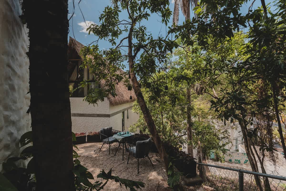 Serene outdoor patio area surrounded by dense tropical jungle foliage, offering a private setting with a small table and two chairs. The property's white-walled, thatched-roof structure is partially visible.