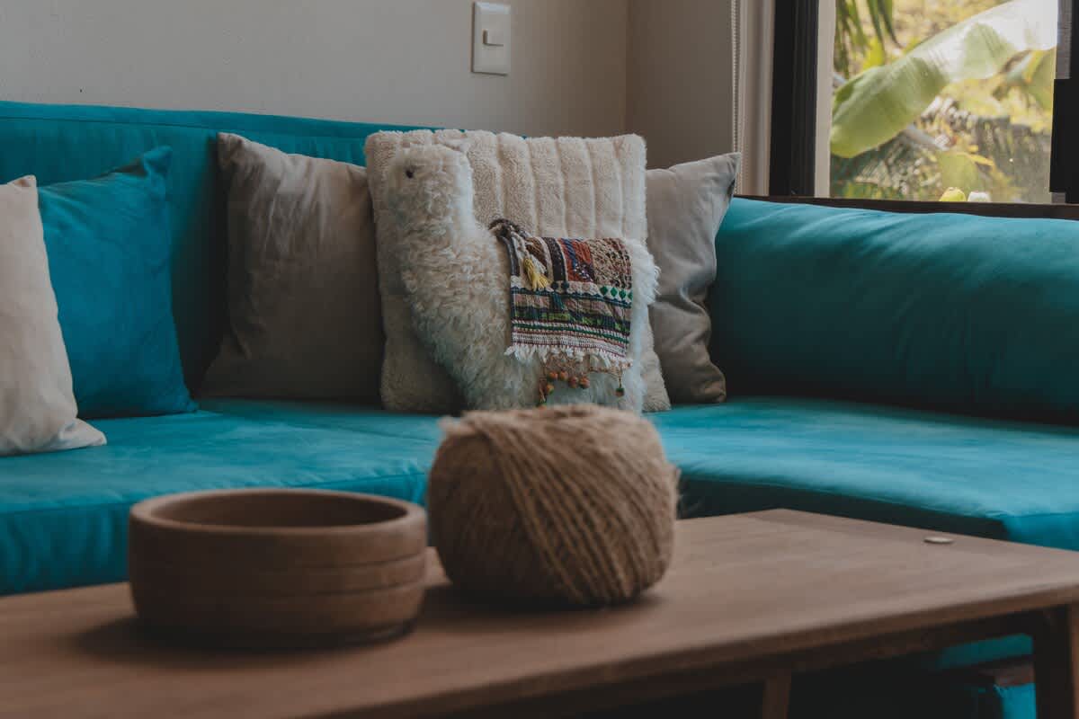 A teal sofa is decorated with a unique llama-shaped pillow and two beige pillows, complemented by a wooden coffee table holding a decorative bowl and a ball of yarn, with natural light from a nearby window.