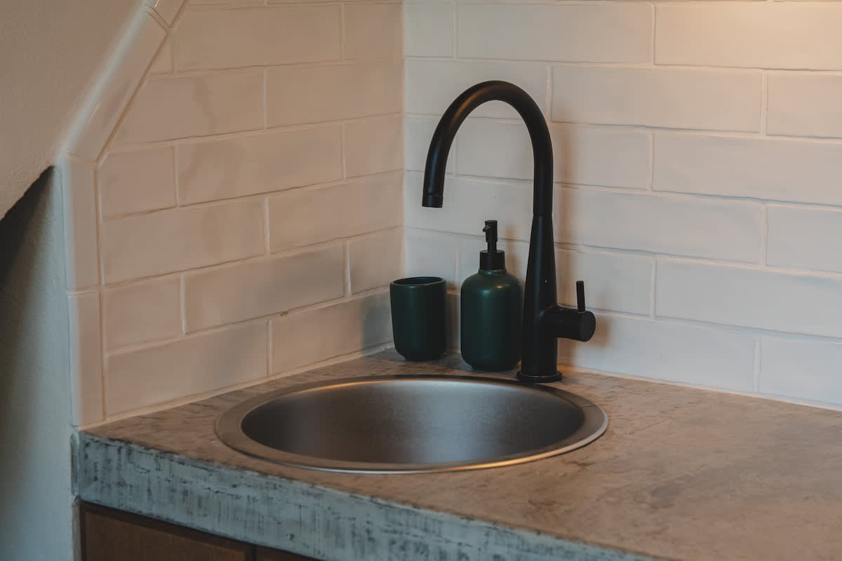 A compact kitchen corner features a stainless steel sink with a modern black gooseneck faucet, a dark green soap dispenser, and a cup, set against a white subway tile backsplash and a concrete countertop.