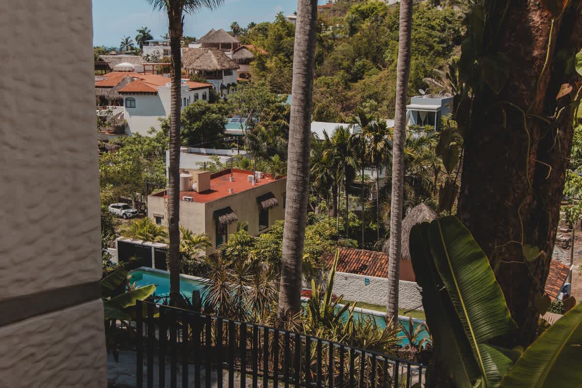 View from a balcony overlooking a rectangular swimming pool and surrounding buildings with red tile roofs, framed by tall palm trees and lush tropical foliage, with a glimpse of the ocean in the distance.