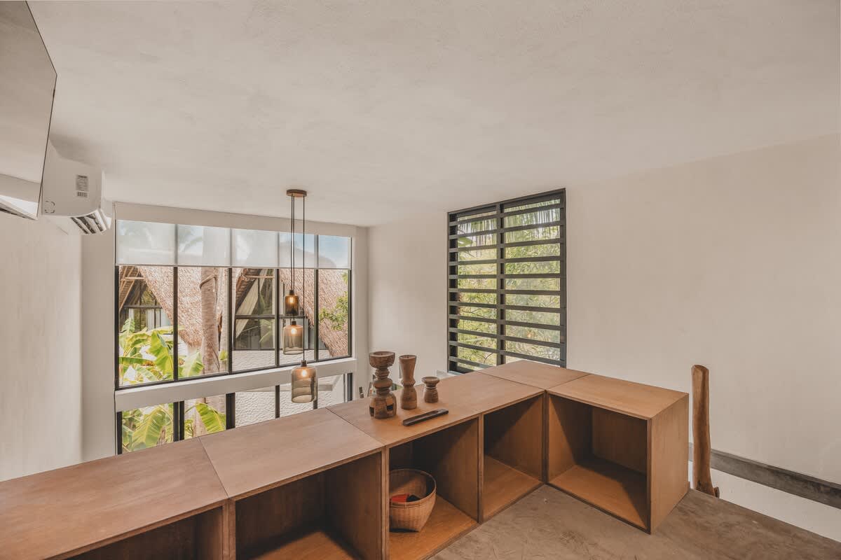 Elevated view of an open common area featuring a long, L-shaped wooden display unit. Large windows offer views of lush tropical foliage and a thatched roof, with modern pendant lights illuminating the space.