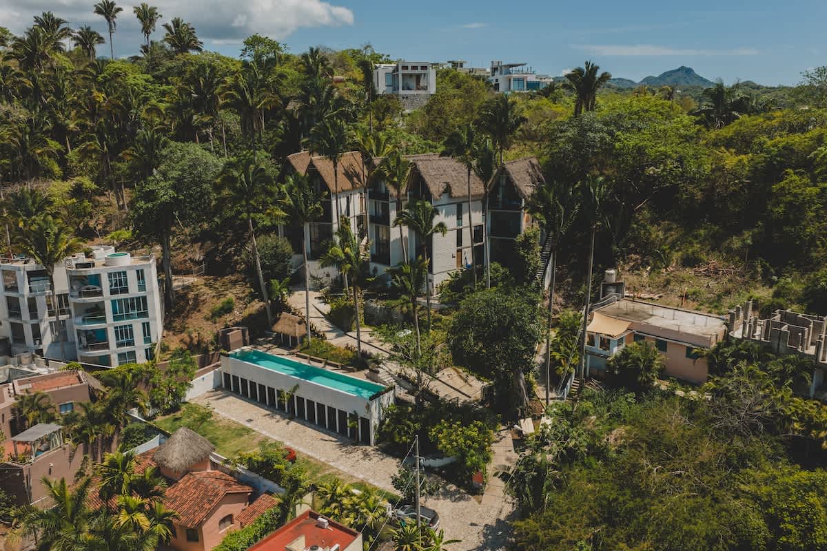 Broad aerial shot of the vacation rental complex, showcasing its distinctive thatched-roof buildings and a long swimming pool, all harmoniously integrated into a vibrant, hilly tropical environment with other nearby homes.