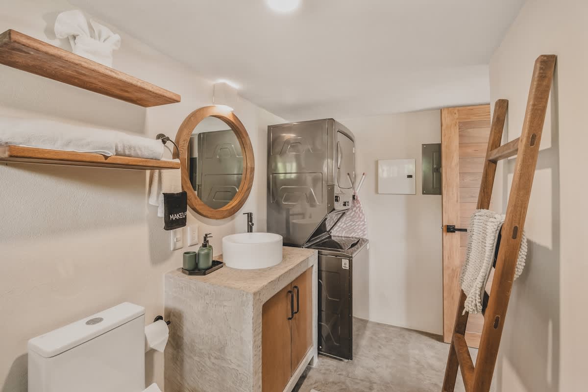 A clean bathroom featuring a concrete vanity with a white vessel sink, a large round wooden mirror, and a stacked washer and dryer unit, with a toilet partially visible.