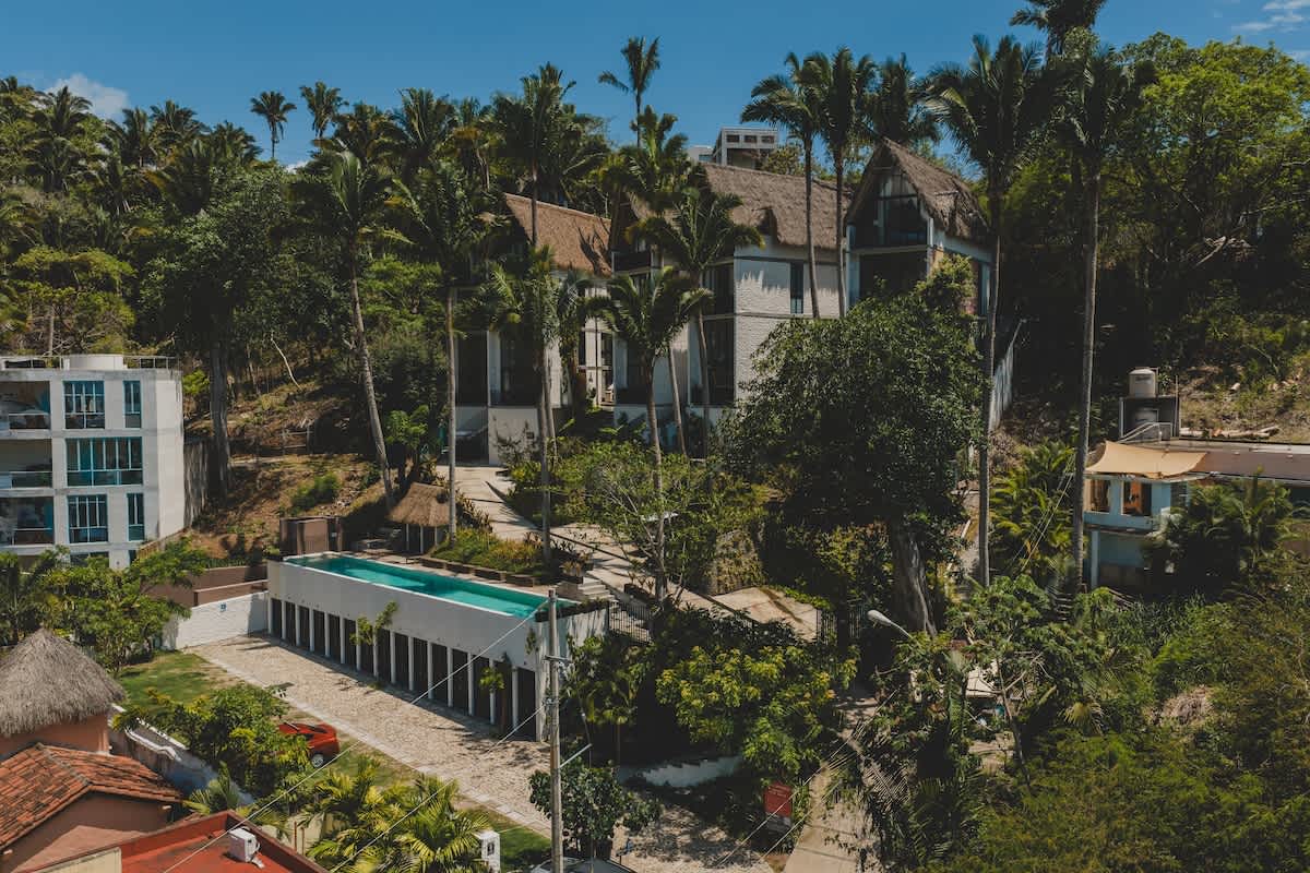 Elevated view of a resort-style property set within a vibrant jungle landscape, highlighting contemporary buildings with unique thatched roofs, a long, narrow swimming pool, and abundant tropical vegetation.