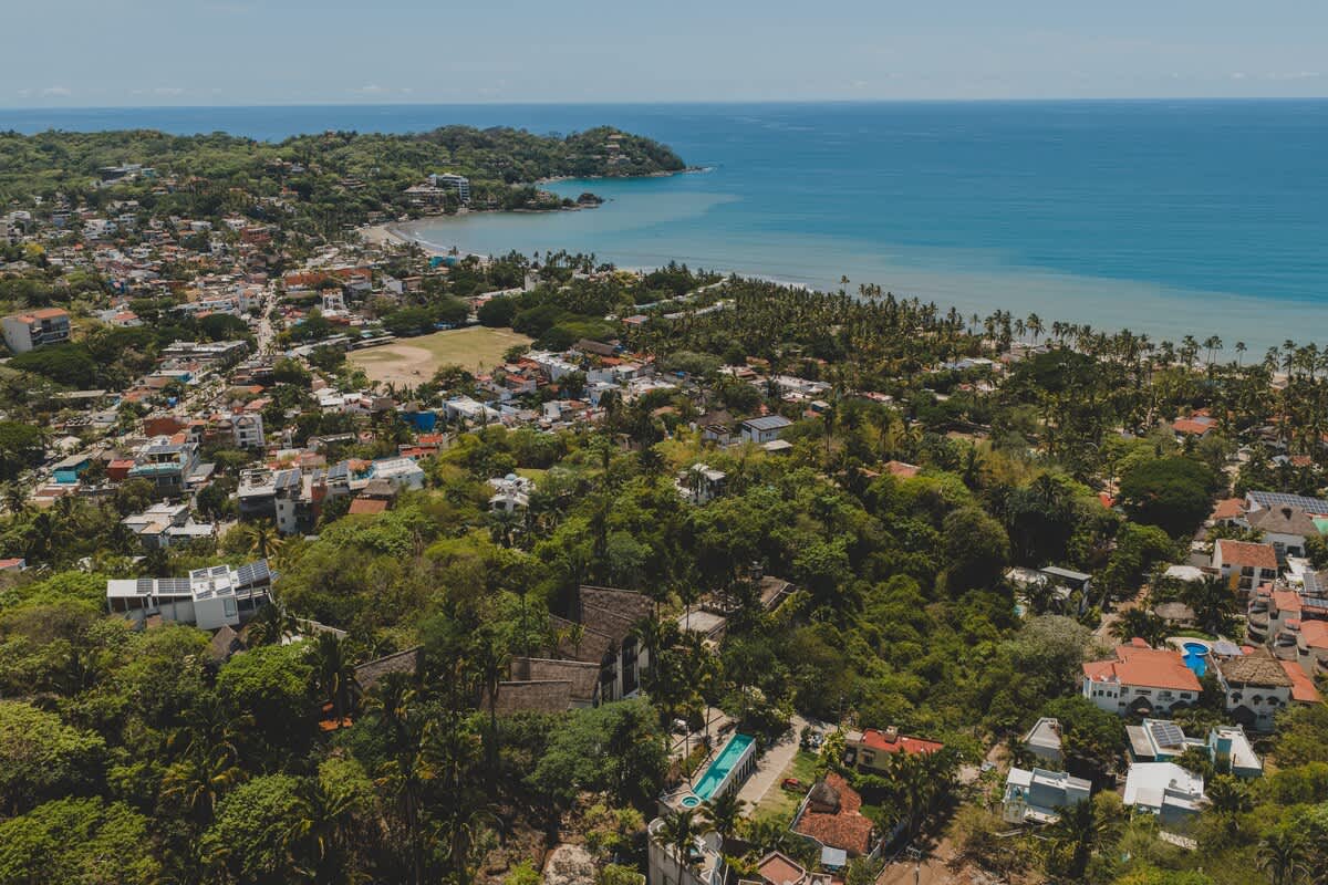 A panoramic aerial view captures a vibrant coastal town with numerous buildings, lush green hills, and a beautiful blue ocean with a sandy beach in the distance under a clear sky. The property is visible amidst the extensive greenery, offering a sense of both seclusion and community.
