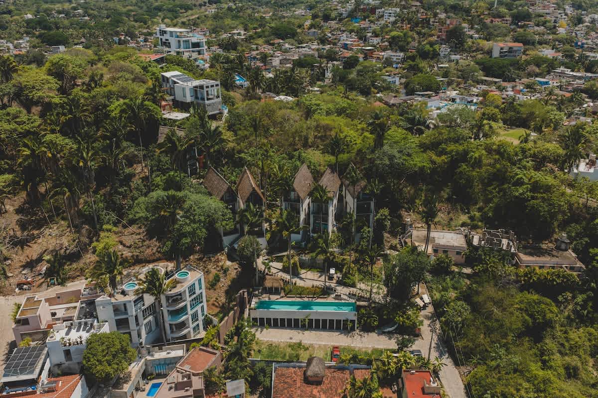 An aerial shot highlights the property's unique architecture with multiple buildings featuring peaked thatched roofs, surrounded by dense tropical trees and a long, rectangular swimming pool. Other residential structures are visible nearby, blending into the lush, green environment.