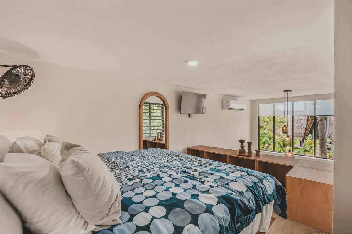 Bright bedroom with a large bed, white pillows, and a blue and white patterned duvet. A wall-mounted TV and AC unit are visible, with a large window overlooking tropical greenery and a wooden console below.