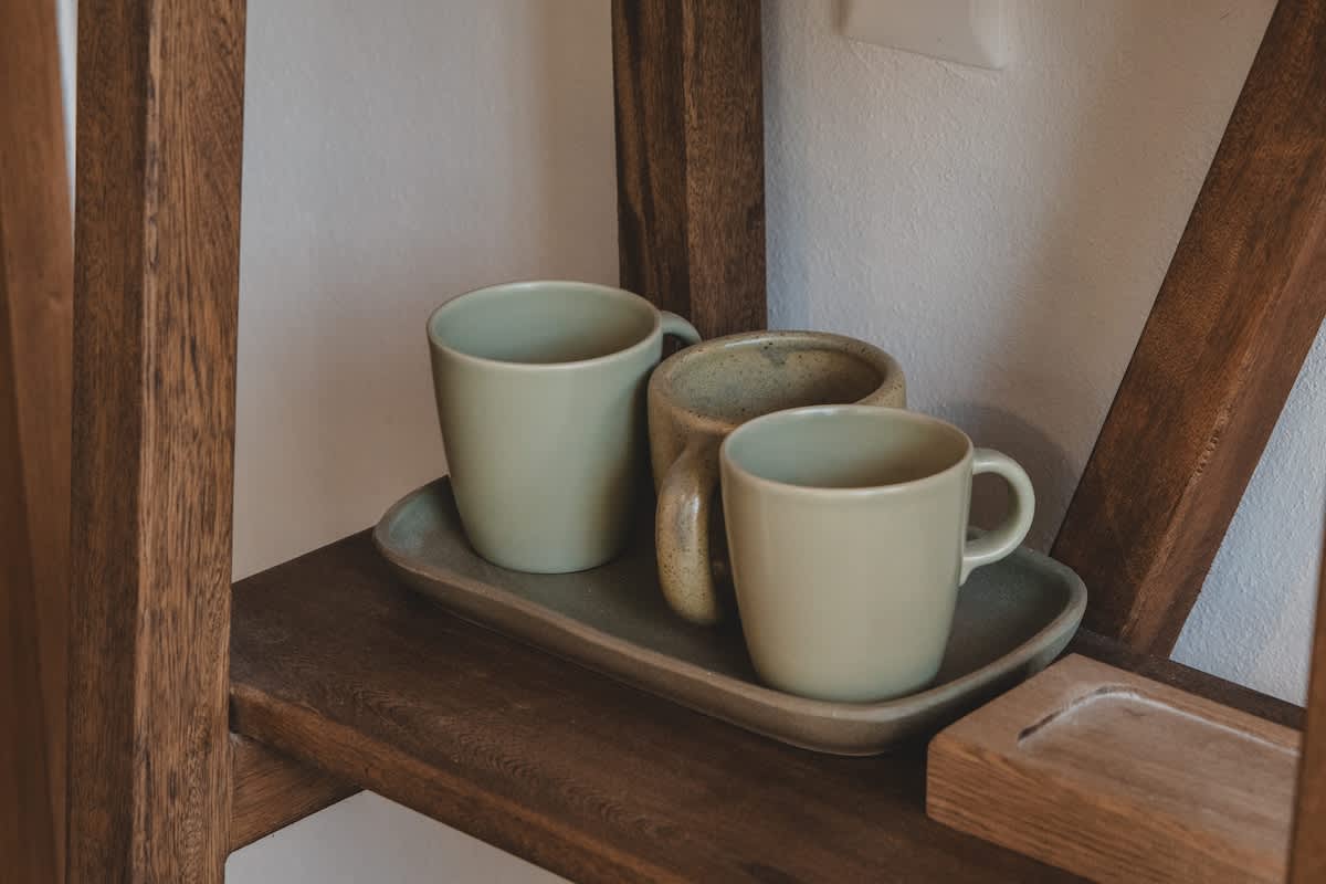 Three light green ceramic mugs are neatly arranged on a rectangular tray, resting on a rustic wooden shelf against a light-colored wall. The natural wood grain of the shelf adds a touch of organic warmth to the decor.