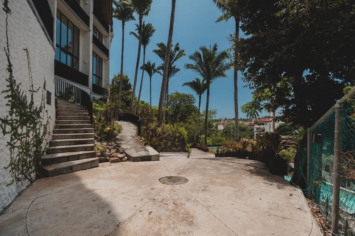A circular concrete turnaround area is flanked by steps leading to a white building, with dense tropical vegetation, tall palm trees, and a chain-link fence visible.