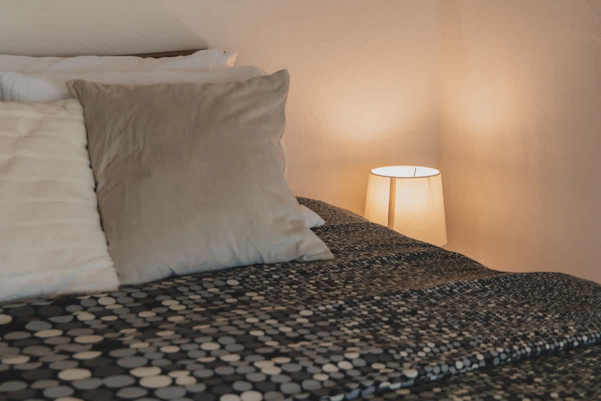 A cozy bedroom features a bed adorned with soft white and light grey pillows, complemented by a dark patterned blanket, and a warm glowing table lamp on a bedside table. The room is illuminated by the lamp's soft light, casting a gentle ambiance on the light pink wall.