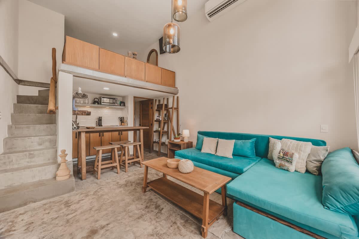 Open-concept living space with a teal sectional sofa, concrete floors, and a wooden breakfast bar with stools. A staircase leads to a mezzanine level, and the area is brightly lit with natural light.