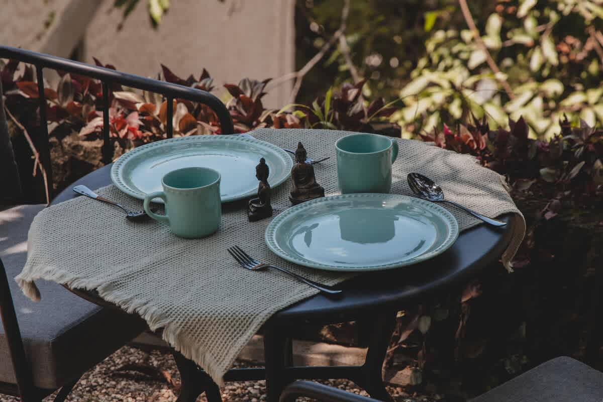 An inviting outdoor dining table is set for two with light blue plates, matching mugs, and silverware on a textured beige placemat, flanked by two small Buddha statues. Lush green and red foliage provides a vibrant, natural backdrop to this serene dining spot.