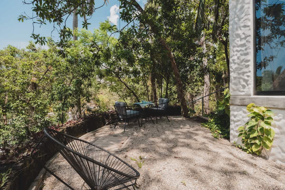 Secluded outdoor patio area with a small round table and two chairs, plus an additional modern black wire chair, set on a gravel surface and surrounded by dense, verdant jungle trees, next to a white building.