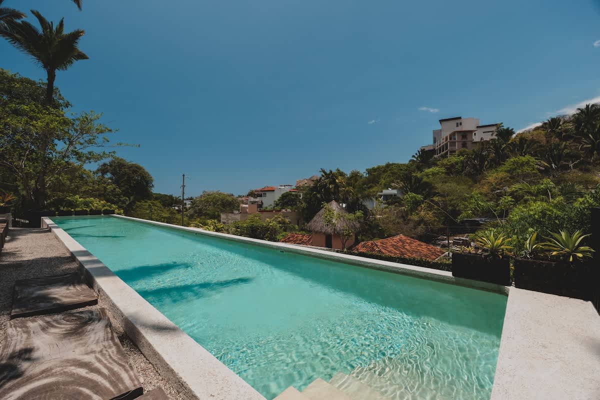 A long, narrow infinity pool with light blue water offers panoramic views of a lush, green hillside dotted with houses under a clear blue sky.