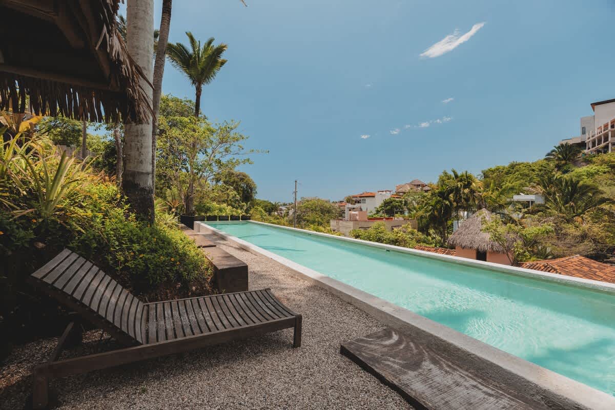 Wooden lounge chairs are positioned alongside a long, narrow swimming pool, offering a serene view of the distant houses and lush tropical landscape under a bright sky.