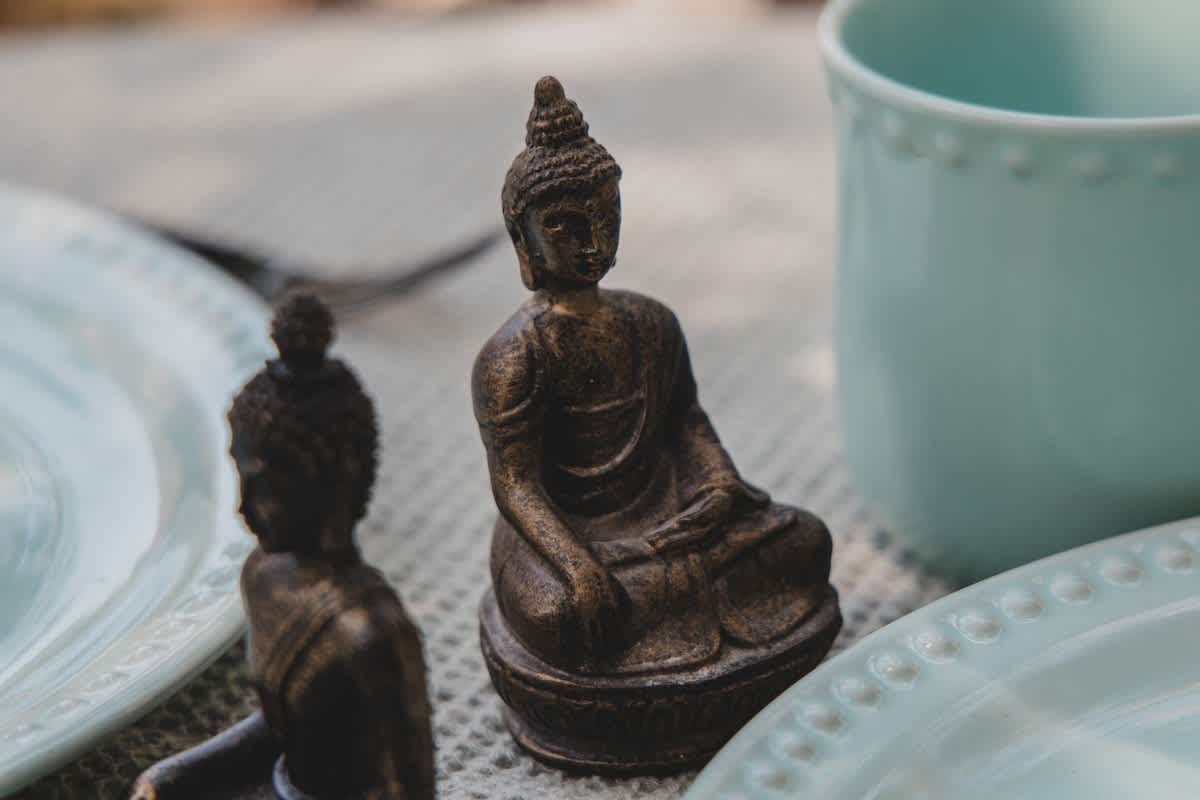 A close-up view captures a small, intricately detailed bronze Buddha statue in a meditative pose, positioned on an outdoor dining table next to a light blue ceramic plate and mug. Another Buddha statue is partially visible in the soft-focus background, adding to the tranquil setting.