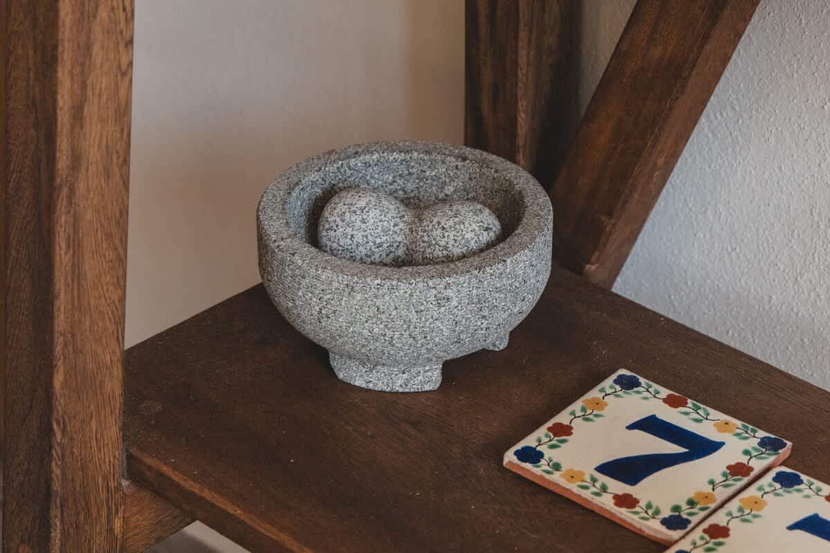 A grey stone mortar and pestle set, featuring two heart-shaped pestles, rests on a rustic wooden shelf next to two decorative ceramic tiles with a blue number '7' and colorful floral patterns. The dark wood of the shelf provides a warm base for the textured items.
