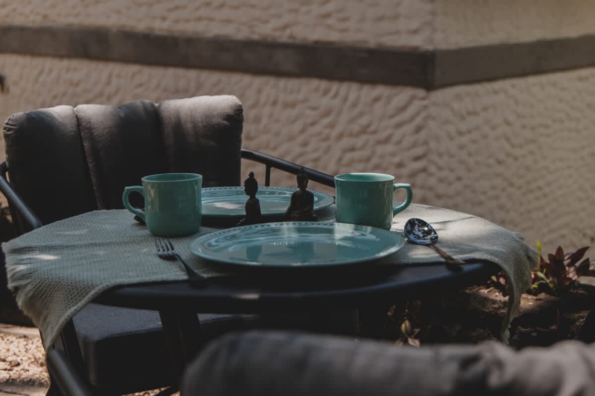 Inviting outdoor table setting with two cushioned chairs, featuring light blue plates, mugs, and small Buddha statues on a round table. The lush green foliage provides a natural, blurred backdrop.