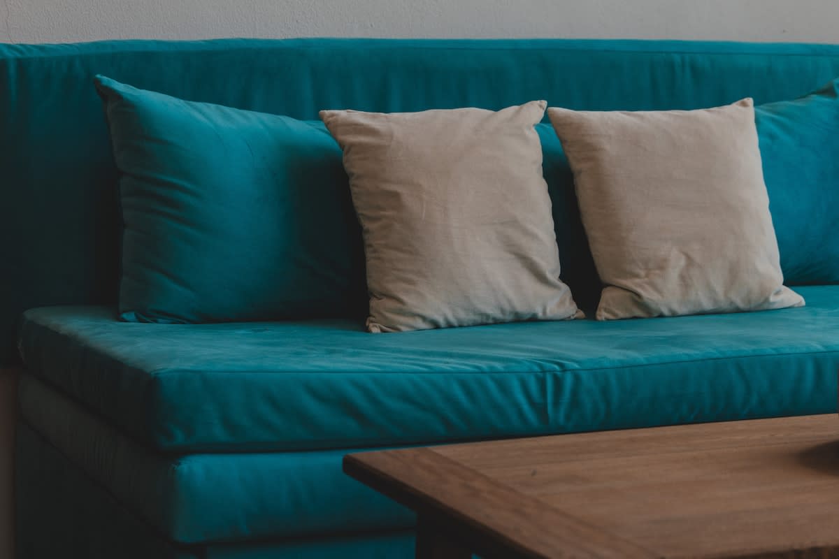 A vibrant teal sofa is adorned with two light beige throw pillows, positioned next to a rustic wooden coffee table, suggesting a comfortable and inviting living space.