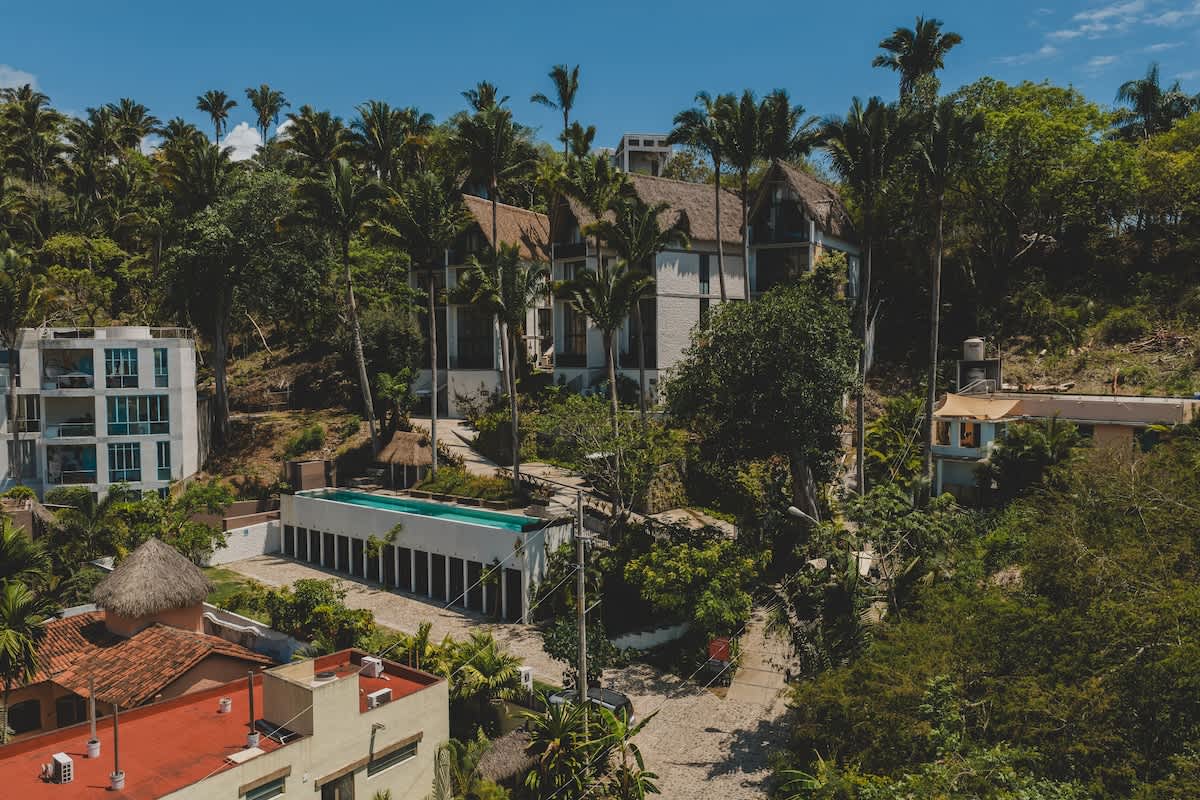 Overhead view showcasing a tropical property with modern buildings featuring thatched roofs, surrounded by dense jungle foliage and tall palm trees, with a prominent rectangular swimming pool in the foreground.