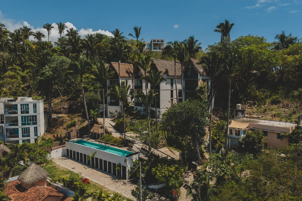 Aerial view of a tropical resort complex nestled in a lush green jungle, featuring several modern buildings with thatched roofs, a long rectangular swimming pool, and surrounding palm trees under a clear blue sky.