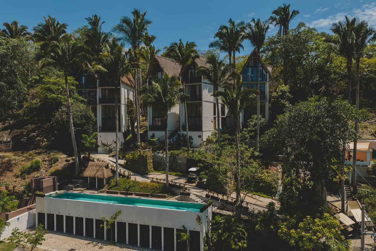 Closer aerial perspective of the property, revealing a series of modern, multi-story buildings with distinctive thatched A-frame roofs, a long, elevated swimming pool, and dense tropical greenery.