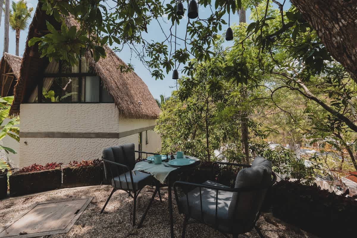 Inviting outdoor patio with a small table and two cushioned chairs, set against a backdrop of lush trees and a distinctive thatched-roof building. Decorative hanging lights add to the ambiance.