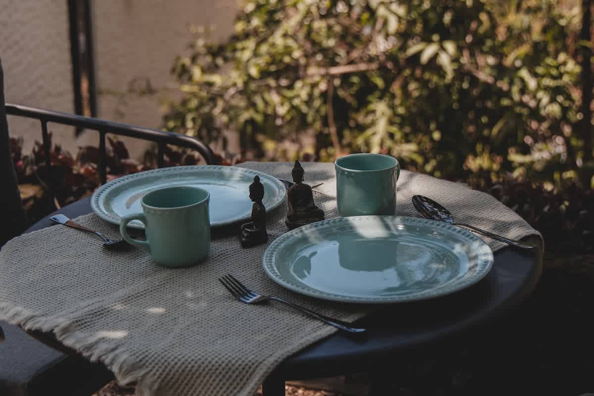 Detailed view of an outdoor dining setup on a small round table, featuring two light blue plates, matching mugs, and cutlery on a textured placemat, flanked by two small Buddha statues.