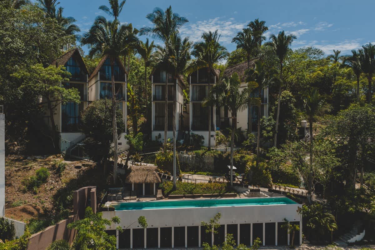 Drone shot capturing the architectural design of the property, featuring several tall, narrow buildings with large windows and thatched roofs, integrated into a steep, jungle-covered hillside with a long pool.