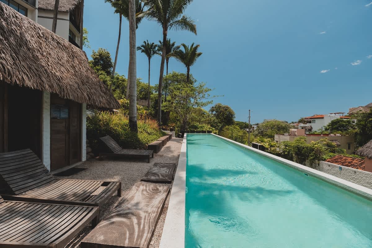 A long, rectangular swimming pool is bordered by wooden lounge chairs and a small thatched-roof cabana, with modern multi-story buildings and tall palm trees in the background.
