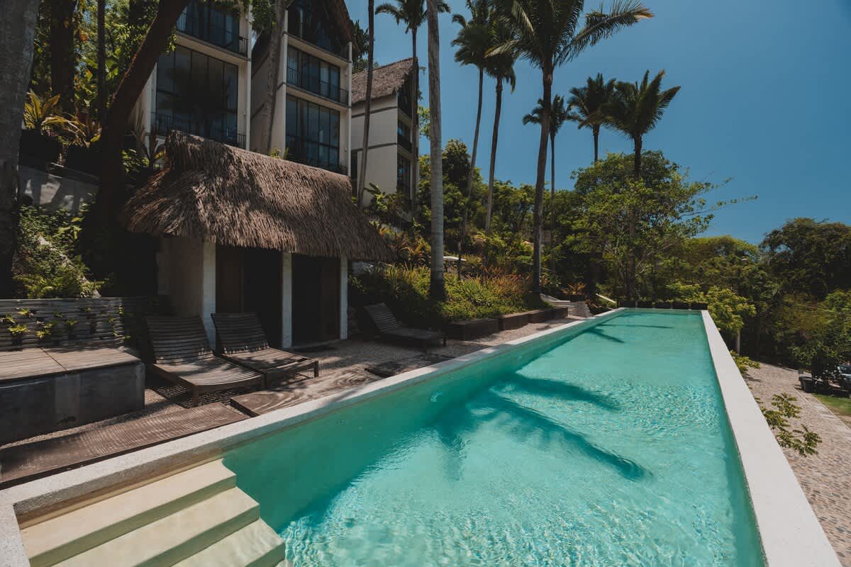 A long, rectangular swimming pool is flanked by wooden lounge chairs and a small hut with a thatched roof, set against a backdrop of modern buildings and tall palm trees.