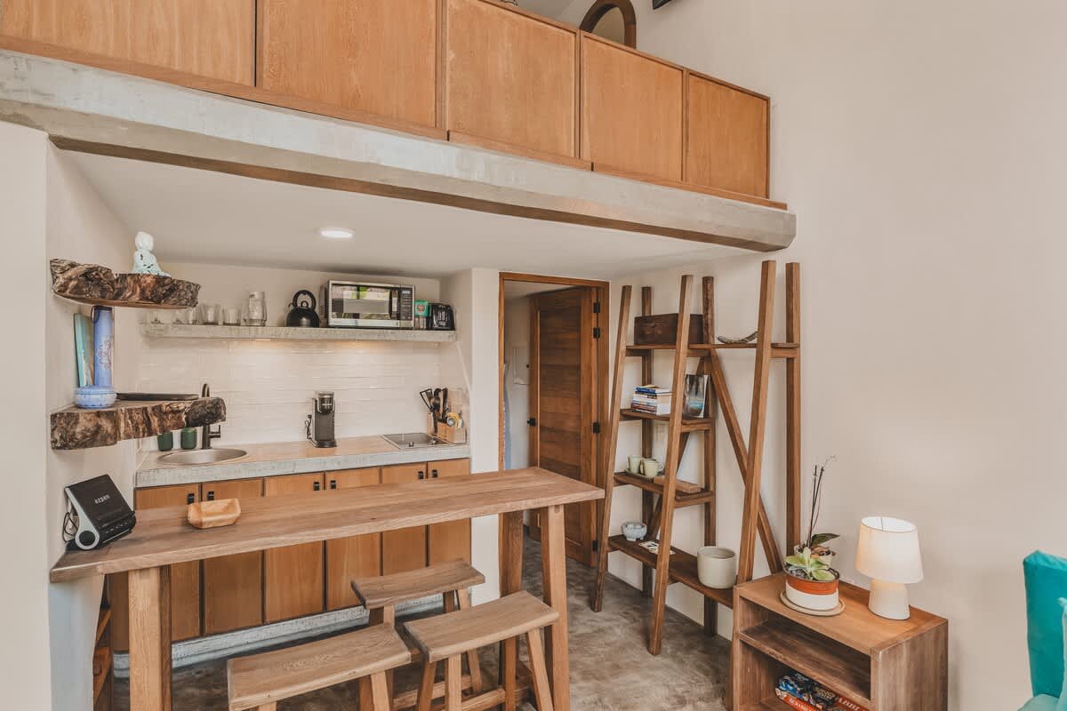 A compact kitchen and dining area featuring a wooden table with stools, concrete countertops, and wooden cabinetry, with a lofted wooden sleeping area visible above.