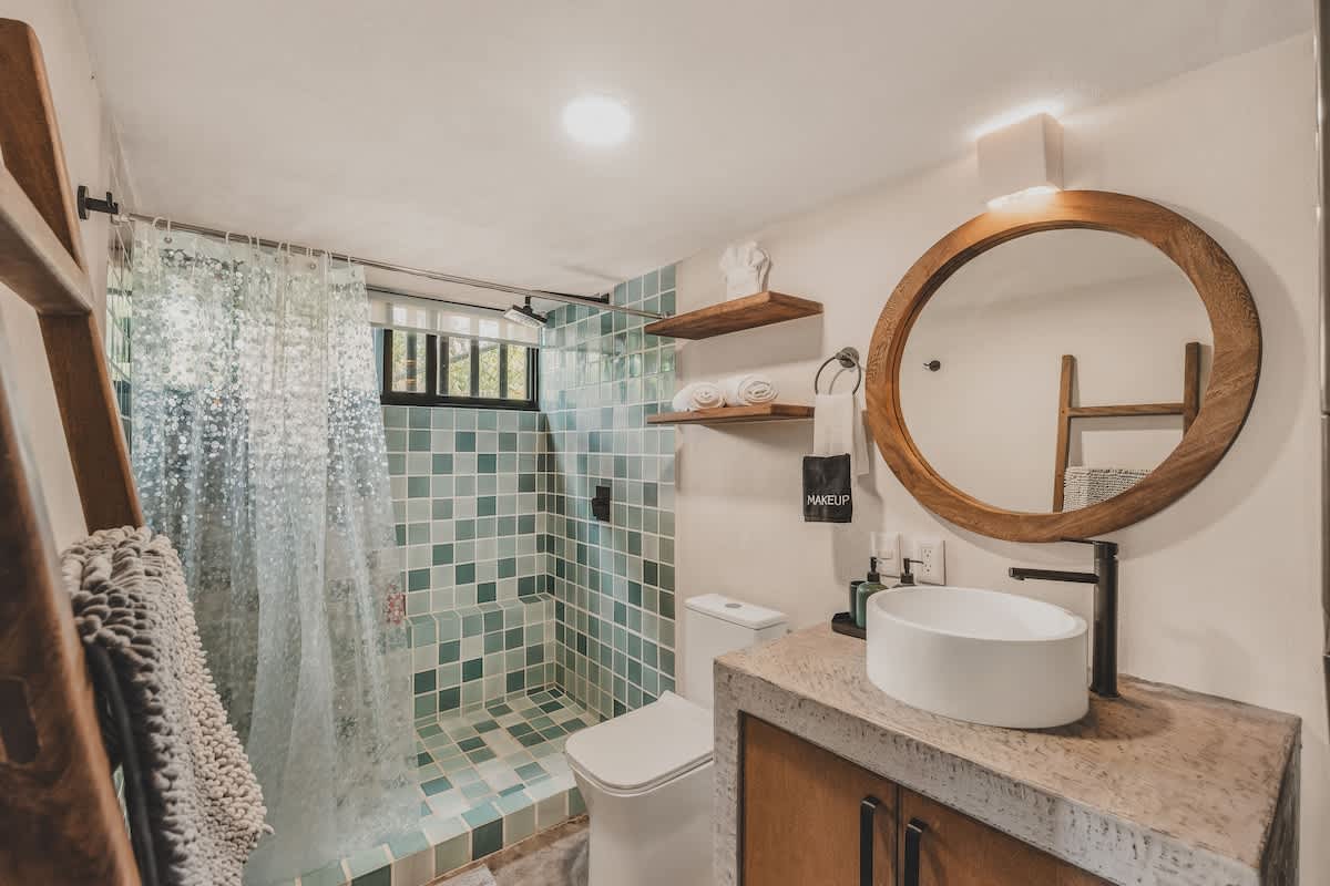 A bright bathroom with a white toilet, concrete vanity, vessel sink, and a shower area tiled with light blue and green square tiles, featuring a clear shower curtain.