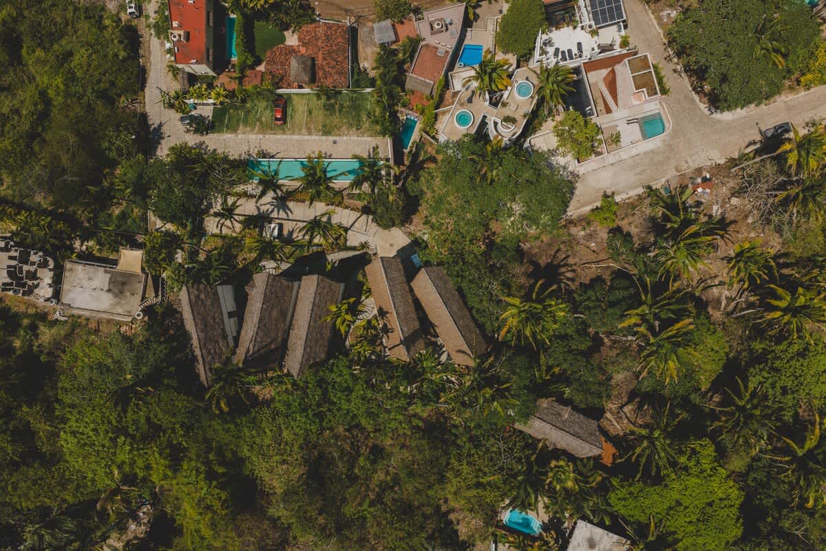An aerial view showcases a cluster of buildings with distinctive thatched roofs nestled within dense green jungle foliage, surrounded by other residential properties and a sparkling swimming pool. The vibrant tropical landscape highlights the property's secluded yet accessible location.