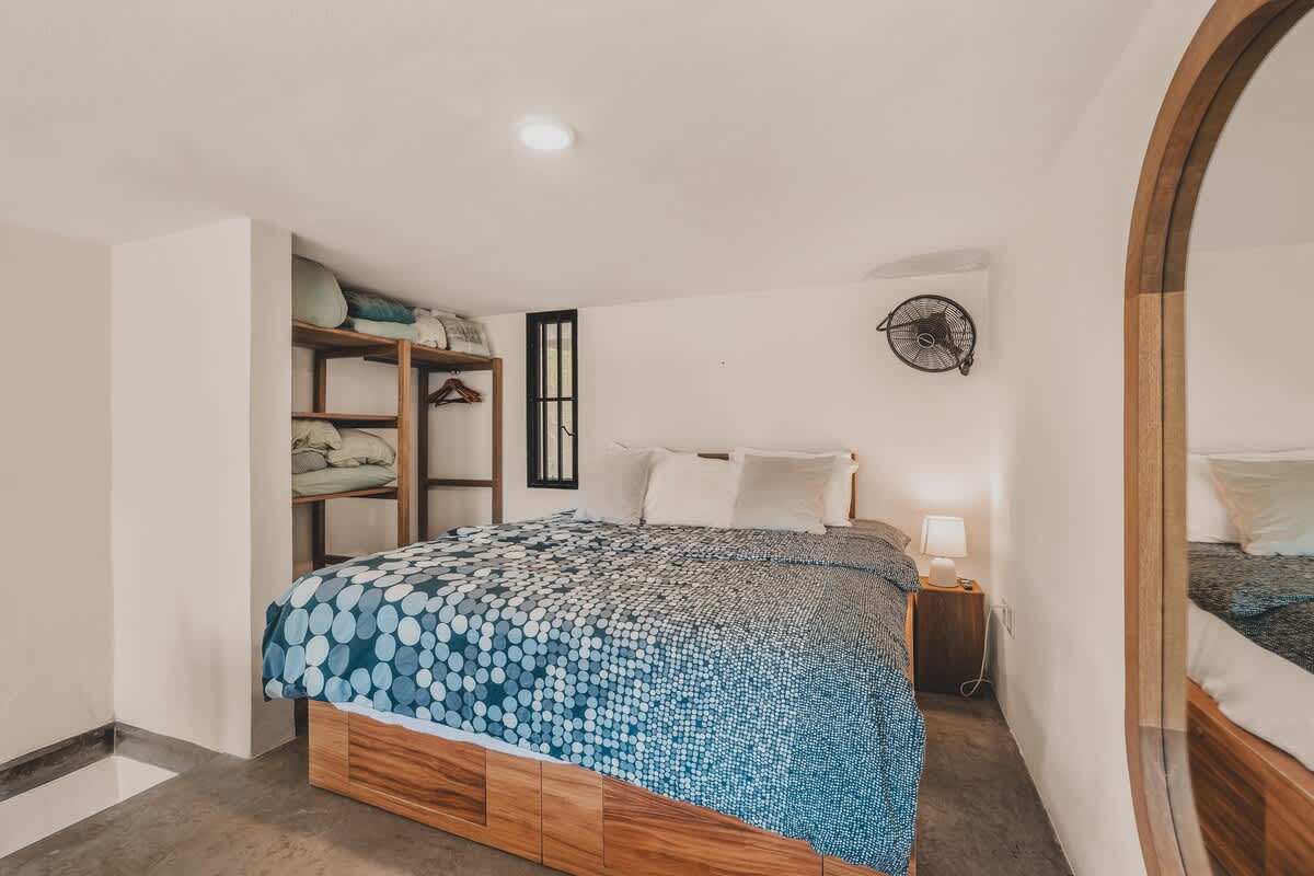 Cozy bedroom featuring a large bed with a blue and white patterned duvet, white pillows, and a wooden base. A built-in wooden shelving unit holds linens, and a wall-mounted fan provides air circulation.