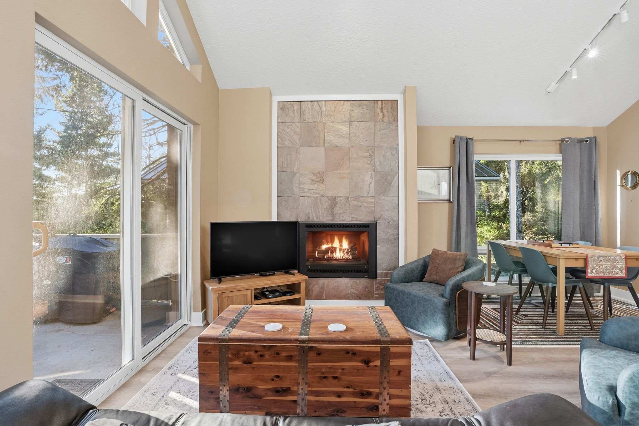 A warm living room featuring light wood-look flooring, a prominent stone-tiled fireplace with a gas insert, and a sliding glass door opening to an outdoor space, with a dining area visible in the background.