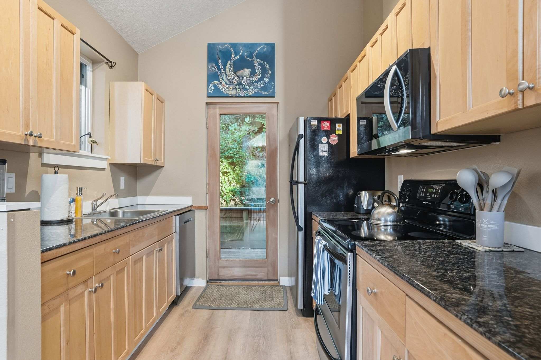 Galley kitchen with light wood cabinets, dark granite countertops, a stainless steel refrigerator, and a black electric stove, featuring a glass-paned door leading to an outdoor area.
