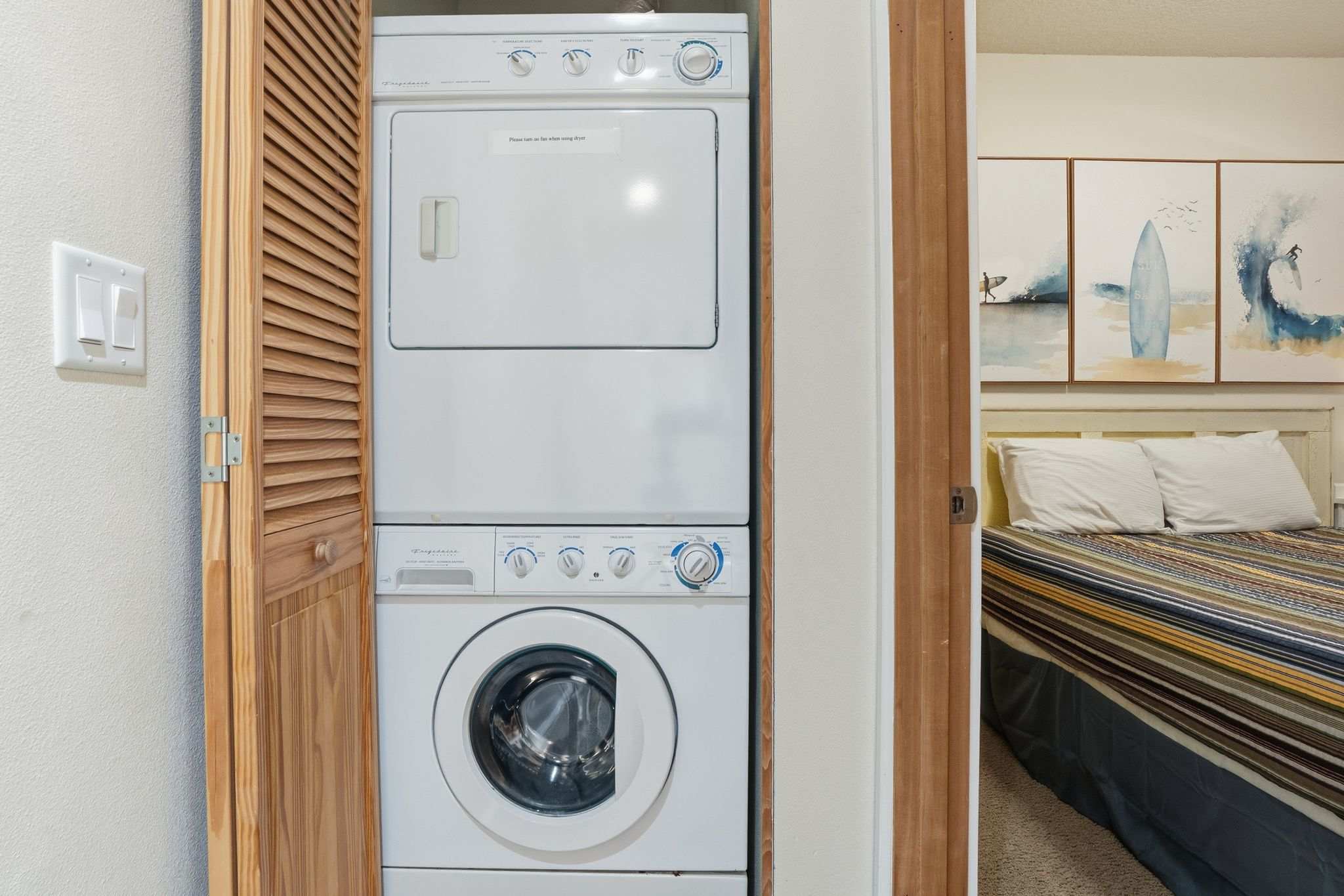 A white stacked Frigidaire washer and dryer unit is neatly tucked into a closet with louvered wooden bi-fold doors. The laundry area is adjacent to a bedroom with a striped bedspread.