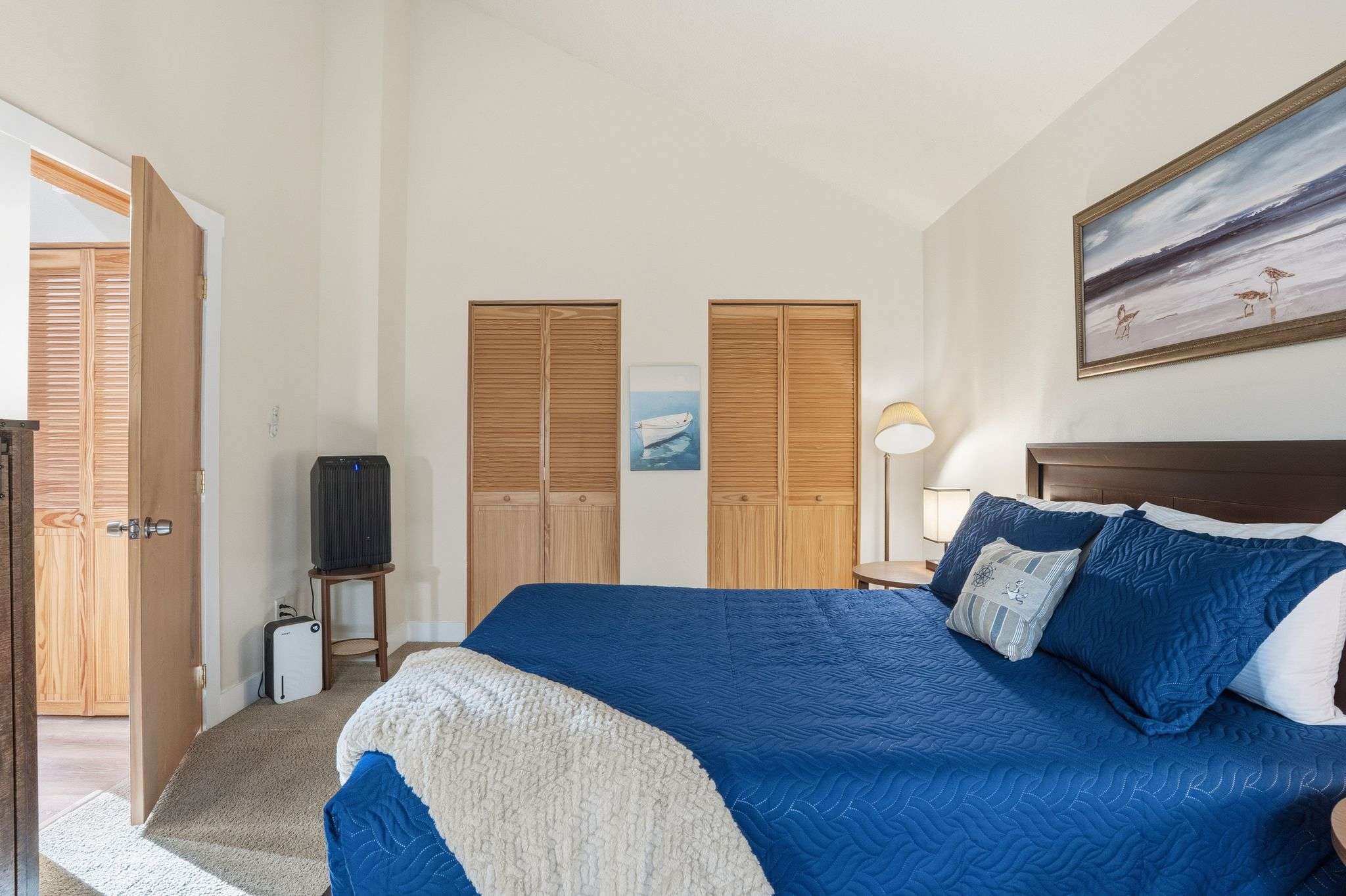 Spacious bedroom with a queen-sized bed, blue quilted bedding, and a dark wood frame, set against light-colored walls with vaulted ceilings. Two louvered closet doors are visible, along with a modern air purifier on a stand.