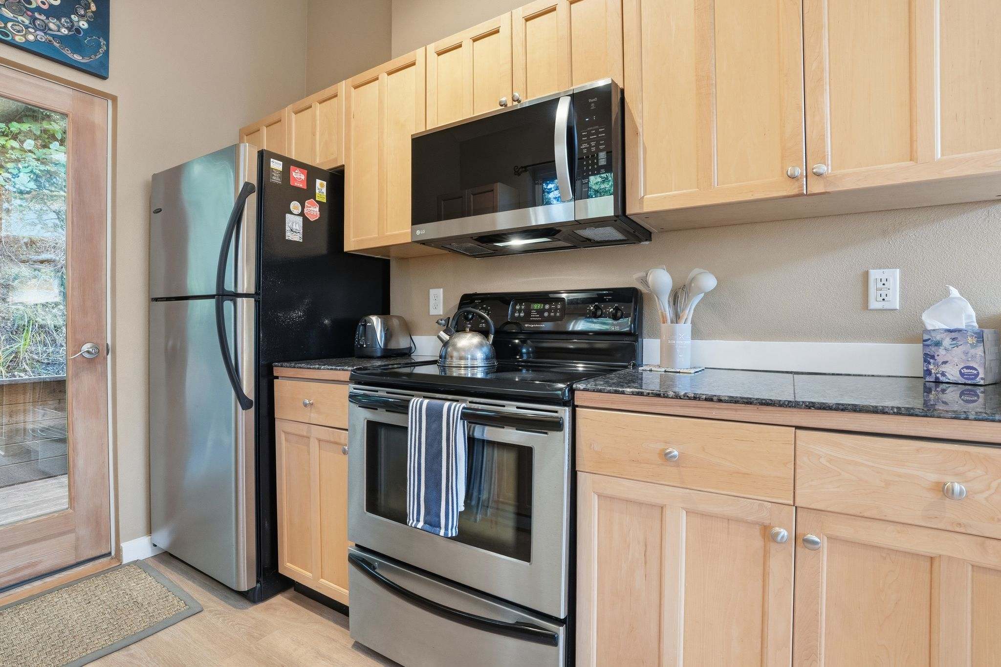 Kitchen area showing a stainless steel refrigerator next to a black electric stove, with light wood cabinets above and dark granite countertops, and a stainless steel microwave overhead.