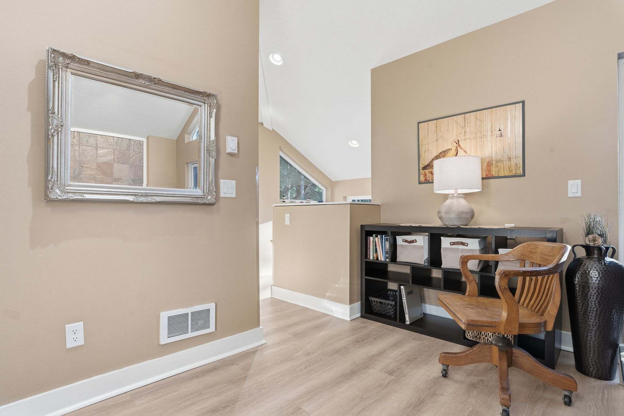 A hallway with light beige walls and light wood flooring features a large ornate silver-framed mirror and a black shelving unit with a wooden desk chair. A table lamp and decorative items adorn the shelving unit, with a window visible in the background.