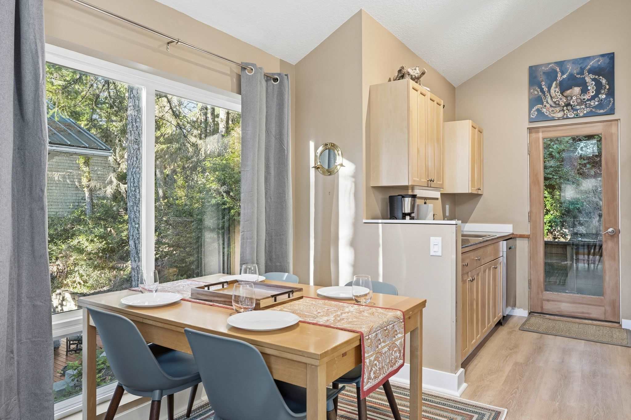 Cozy dining area with a light wood table and blue chairs, positioned next to a large window offering views of lush greenery. A compact kitchen nook with light wood cabinets and a coffee maker is visible in the background.