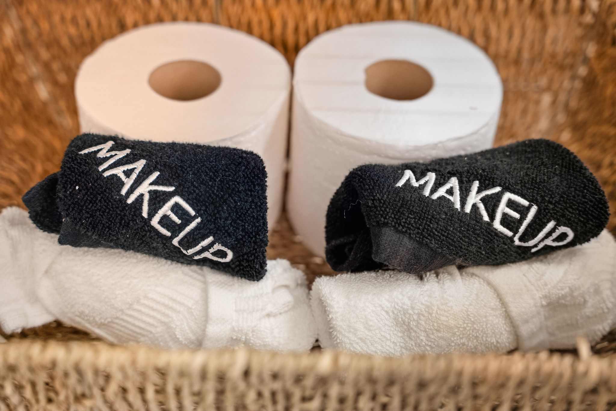 A close-up shows a woven basket holding two rolls of toilet paper, two black makeup remover towels, and several white hand towels, neatly arranged for guest use.